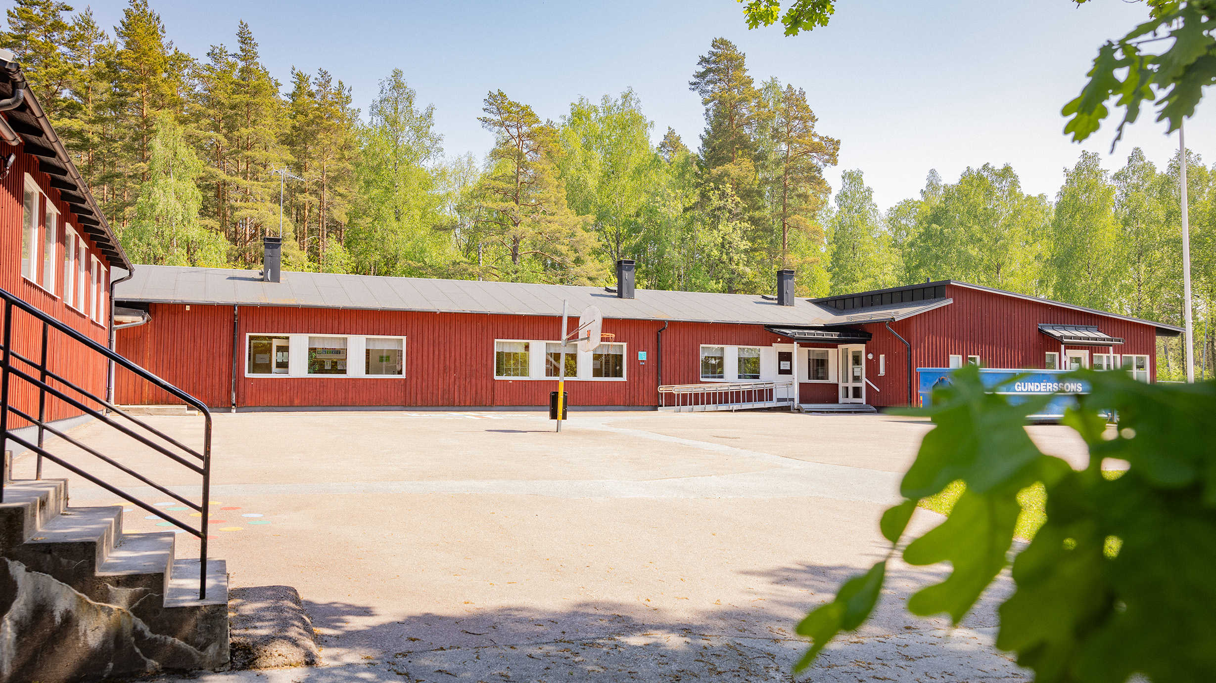 Röd träskola med grusplan i förgrunden och bakomliggande skog.